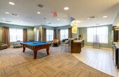 Located in Arlington, MA, this single-floor Media & Game Room is a well-appointed communal space designed for both recreation and relaxation. The open main area is anchored by a full-size billiards table set atop a distinctive herringbone-patterned carpet, surrounded by comfortable seating and bathed in natural light from large windows dressed with elegant draping curtains. Soft blue-gray walls and recessed lighting lend the space a calm, welcoming ambiance throughout.

A dedicated media lounge area features a large wall-mounted flat-screen television set against rich dark wood cabinetry, complemented by a mix of accent chairs and a small round table — ideal for intimate viewing or casual gatherings. Track lighting and framed artwork add a refined, curated aesthetic to this zone.

A semi-private nook, accessible through a modern sliding barn door, offers additional seating alongside natural light and tall decorative plants, making it well-suited for quieter activities or small group use.

The kitchenette is fully equipped with dark cabinetry, a sink, microwave, under-counter refrigerator, and a coffee station, all finished with light tile flooring and a decorative tile backsplash. Together, these distinct zones create a multifunctional space perfect for socializing, entertainment, and everyday leisure.