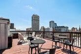This rooftop deck delivers an open-air experience elevated above the surrounding cityscape, offering sweeping panoramic views of the downtown skyline and beyond. The warm-toned hardwood decking spans a generous footprint and is thoughtfully organized into several distinct zones, each serving a different function while contributing to the deck's overall versatility.

A covered pavilion anchors one section of the deck, featuring a metal roof supported by wooden posts and flanked by sheer curtain panels. This shaded area provides shelter from the elements and is outfitted with bistro-style tables and chairs, making it well-suited for casual dining or conversation. Outdoor grills positioned nearby add functional value for gatherings.

Adjacent open-deck areas are furnished with classic white Adirondack chairs arranged around a central fire pit, creating a relaxed lounge setting beneath the open sky. Colorful outdoor rugs define the seating areas and add visual warmth. Raised planter boxes with ornamental grasses and string lights contribute a polished, inviting aesthetic.

A railing system encloses the full perimeter of the deck, ensuring safety while maintaining unobstructed sightlines to the surrounding skyline. The combination of covered and open spaces, diverse seating arrangements, and commanding urban views makes this rooftop deck a dynamic and well-appointed outdoor destination.