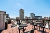 This rooftop deck delivers an open-air experience elevated above the surrounding cityscape, offering sweeping panoramic views of the downtown skyline and beyond. The warm-toned hardwood decking spans a generous footprint and is thoughtfully organized into several distinct zones, each serving a different function while contributing to the deck's overall versatility.

A covered pavilion anchors one section of the deck, featuring a metal roof supported by wooden posts and flanked by sheer curtain panels. This shaded area provides shelter from the elements and is outfitted with bistro-style tables and chairs, making it well-suited for casual dining or conversation. Outdoor grills positioned nearby add functional value for gatherings.

Adjacent open-deck areas are furnished with classic white Adirondack chairs arranged around a central fire pit, creating a relaxed lounge setting beneath the open sky. Colorful outdoor rugs define the seating areas and add visual warmth. Raised planter boxes with ornamental grasses and string lights contribute a polished, inviting aesthetic.

A railing system encloses the full perimeter of the deck, ensuring safety while maintaining unobstructed sightlines to the surrounding skyline. The combination of covered and open spaces, diverse seating arrangements, and commanding urban views makes this rooftop deck a dynamic and well-appointed outdoor destination.
