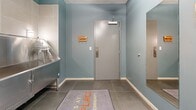 Located at 1507 Fifth Ave N in Nashville, Tennessee, this dedicated pet wash facility offers a well-designed, single-floor space built specifically for pet grooming and care. The room is finished with cool blue-gray walls and large-format gray tile flooring, giving the space a clean, practical, and welcoming aesthetic.

The centerpiece of the space is a professional-grade stainless steel pet bathing tub, complete with side access panels, an integrated rinsing attachment, a grooming restraint loop, and a step platform for easy pet entry. Wall-mounted sconce lighting flanks the tub area, providing ample illumination. Adjacent to the tub is a stainless steel prep table with a lower shelf, ideal for organizing grooming supplies, set against floor-to-ceiling neutral tile walls with a conveniently placed floor-level water spigot.

The main area features a skylight that brings in natural overhead light, enhancing the open feel of the space. A decorative gallery wall showcases dog-themed framed artwork and wooden signs, adding warmth and personality. A wooden ladder-style shelving unit provides additional storage. A floor drain in the center of the room ensures easy cleanup. The entryway includes a welcoming mat, wall-mounted hooks for leashes and accessories, and a full-length mirror, making this an efficient and thoughtfully appointed pet care amenity.