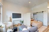 This thoughtfully designed studio in Haverhill, MA, makes the most of its efficiently organized single-floor layout. A welcoming hallway leads into the heart of the unit, flanked by two closets and a utility room, providing ample storage solutions rarely found in a studio format. The open-plan main living area seamlessly combines the living room, dining space, and kitchen into one bright, airy environment. Soaring ceilings amplify the sense of space, while two large black-framed windows flood the room with natural light and frame sweeping views of lush treetops and the surrounding Haverhill landscape. Light-toned hardwood-style flooring flows throughout, enhancing the clean, contemporary aesthetic. The kitchen is outfitted with modern wood-toned and white cabinetry, a white subway tile backsplash, a sleek countertop, and a full suite of stainless steel appliances. A sliding barn door opens to a dedicated sleeping area, offering a clever touch of privacy within the open layout. The full bathroom features a glass-enclosed walk-in shower with floor-to-ceiling white tile, black hardware accents, a floating vanity, and a frameless mirror. In-unit laundry adds convenience, completing this well-appointed, modern studio.