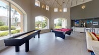 This single-story community clubhouse is designed to offer a diverse range of amenities within a cohesive, modern layout. The centerpiece is a dramatic game room boasting soaring vaulted ceilings with exposed truss detailing, arched windows that flood the space with natural light, and views of the surrounding tropical landscaping. A full-size billiards table and shuffleboard table make it a premier destination for recreation and social gatherings.

Adjacent to the game room is a stylish lounge area featuring contemporary seating, a wall-mounted flat-screen television, and bold red and gray geometric wall art that adds a vibrant, modern touch. The connected business center offers leather seating, a workstation with computer and printer access, and a similarly striking accent wall.

A bright, windowed sunroom provides a comfortable retreat with cushioned lounge seating, a ceiling fan, and panoramic views of mature tropical greenery. The covered outdoor patio area is equipped with a built-in grilling station, large dining tables, and bar-height seating — ideal for outdoor entertaining. Pendant lighting throughout adds warmth and character to each area of this well-rounded clubhouse facility.