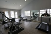 This fitness center offers a bright, open, and inviting environment designed to support a comprehensive workout routine. The space is defined by its impressive vaulted ceilings with recessed lighting, which contribute to an airy, spacious atmosphere. Light-toned hardwood-style flooring runs throughout, providing a clean and modern aesthetic that complements the neutral gray walls.

Natural light streams in generously through multiple multi-pane windows positioned along the walls, while a set of French doors opens directly to an outdoor area, offering views of what appears to be a pool and surrounding greenery. This seamless indoor-outdoor connection enhances the overall ambiance of the space.

The fitness center is outfitted with a well-rounded selection of cardio equipment, including multiple treadmills, an elliptical trainer, a stationary spin bike, and a rowing machine. A large wall-mounted mirror spans a significant portion of one wall, making the space feel even more expansive and allowing users to monitor their form during exercise. Protective rubber mats are placed beneath the equipment to preserve the flooring.

Additional amenities include a small cabinetry unit, likely used for storage or refreshments, and ample open floor space for stretching or mat-based workouts. The overall design is clean, functional, and well-maintained, making this fitness center a practical and pleasant amenity.