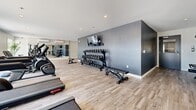 Located at 17 Place De La Triade in Pointe-Claire, Quebec, this fitness facility offers a generously sized, single-floor exercise room designed to accommodate a comprehensive range of workout activities. The space features light wood-effect flooring throughout, which contrasts cleanly with a combination of crisp white and bold charcoal-grey accent walls, giving the gym a modern, professional aesthetic.

The cardio zone is well-appointed, housing multiple Star Trac treadmills, an elliptical trainer, stationary bikes, and a rowing machine — all arranged to take advantage of the large windows along one wall, which flood the space with natural light and provide views of the outdoor surroundings. A dedicated strength training area features a multi-station cable machine system and additional cable apparatus, offering versatile resistance training options.

A clearly defined free weights section lines the charcoal accent wall, with a full rack of dumbbells organized by weight, accompanied by a flat workout bench and a wall-mounted flat-screen TV. Recessed ceiling lighting provides consistent illumination throughout, while a wall-mounted air conditioning unit ensures a comfortable training environment year-round. A separate utility area with direct access is also present. The overall layout is open and thoughtfully organized, supporting both individual and group fitness routines with ease.