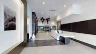 This single-floor lobby presents a contemporary architectural design characterized by a clean, open layout and a refined palette of contrasting dark and light tones. The flooring transitions between large-format light stone tiles and darker tiled sections, visually defining different zones within the space. A striking white and dark sculptural reception desk serves as the focal point, its curved and angular forms creating a bold statement upon entry.

Overhead, a series of distinctive cross-shaped pendant light fixtures hang at varying heights, adding an artistic dimension to the high ceiling above. Recessed lighting throughout ensures the space is evenly and warmly illuminated. Dark wood-paneled wall accents contrast sharply against crisp white walls, lending depth and sophistication to the interior.

Toward the rear of the lobby, glass partitions reveal an adjacent workspace or lounge area, maintaining visual connectivity while defining separate functional zones. A minimalist white decorative vase adds a subtle organic element near the dark accent wall. The overall ambiance is polished and professional, combining modern design sensibilities with practical spatial planning to create a welcoming yet impressive entrance environment.