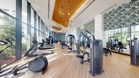 This expansive fitness center is designed with both functionality and aesthetics in mind, offering a well-appointed space for a full range of workout activities. The facility is organized into distinct zones, including a dedicated cycling studio enclosed by sleek glass-panel partitions, a cardio area lined with treadmills and elliptical machines, and a strength training section equipped with a comprehensive selection of cable machines, weight benches, and resistance equipment.

Architecturally, the space is defined by its soaring ceilings adorned with sculptural, organically shaped wood panels finished in warm tones, complemented by decorative starburst pendant lights that add a refined, boutique-hotel ambiance. Floor-to-ceiling glazed walls run the length of the facility, flooding the interior with abundant natural light and providing open views of lush landscaping and surrounding greenery. The light wood-tone flooring enhances the bright, airy atmosphere throughout.

Accent walls featuring bold tropical leaf-pattern wallcoverings introduce a vibrant, resort-inspired character, while large wall mirrors in the strength zone expand the sense of space. A relaxed lounge corner with cushioned seating and a mounted television offers a comfortable recovery area. The overall design balances high-performance fitness functionality with an elevated, visually engaging aesthetic.