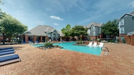 Located at 11212 Westpark Dr in Houston, Texas, this single-level outdoor amenity space is centered around a generously sized resort-style swimming pool with crystal-clear blue water. The pool area is framed by elegant brick-paved decking that extends throughout the space, providing ample room for lounging and recreation. Decorative landscaped islands within and around the pool feature native agave plants, ornamental grasses, and natural stone accents, adding a polished, resort-like aesthetic.

A water feature cascades into the pool, enhancing the ambiance with both visual appeal and soothing sound. Surrounding the pool are multiple navy blue chaise lounges and sun chairs positioned to take advantage of the open sky and natural light. Mature shade trees are thoughtfully integrated throughout the grounds, offering relief from the Texas sun.

The clubhouse building anchors the space architecturally, featuring a classic combination of brick masonry, pitched rooflines, and covered colonnade-style porches. A separate pergola structure with brick columns and white lattice panels provides a shaded seating area with additional outdoor furniture. The entire area is enclosed by a wrought-iron security fence, ensuring privacy and safety. The overall atmosphere is inviting, well-maintained, and designed for outdoor leisure.