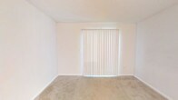 This single-story space presents a straightforward, functional layout characterized by its clean and neutral aesthetic. The room visible features wall-to-wall beige carpeting that extends across the floor, providing a soft and understated foundation. The walls are finished in a crisp white, contributing to a bright and open atmosphere. A textured white ceiling adds a subtle dimensional quality overhead.

A prominent feature of the room is the full-width vertical blind installation, which covers what appears to be a sliding glass door or large window. The blinds allow diffused natural light to filter into the space, creating a softly illuminated environment while maintaining privacy. The white color of the blinds complements the overall neutral tone of the room seamlessly.

White baseboards frame the perimeter of the room, adding a clean finishing detail that ties together the walls and flooring. The space is entirely vacant, allowing for a clear assessment of its dimensions and layout potential. The overall ambiance is calm and minimal, offering a blank canvas well-suited for a variety of furnishing and decorating approaches.