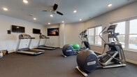 Located in Wisconsin Dells, WI, this dedicated fitness center offers a comprehensive workout facility designed to accommodate a variety of exercise routines. The single-floor space features durable dark carpet flooring throughout, providing a stable and comfortable surface for physical activity.

The room is outfitted with a strong selection of Precor cardio equipment, including elliptical trainers, treadmills, and a stationary bike, giving users multiple options for aerobic exercise. Additional fitness accessories include a set of free weights, green stability balls, and ample open floor space for stretching or bodyweight workouts.

The facility is well-lit by recessed ceiling lights, complemented by a ceiling fan that promotes air circulation during intense sessions. Multiple windows along the walls allow natural light to filter in, creating a bright and inviting workout environment with views of the surrounding Wisconsin Dells area.

Entertainment is provided by multiple wall-mounted flat-screen televisions, including a large display featuring a motivational landscape photograph, keeping users engaged during their workouts. Two access doors provide convenient entry and exit points. The clean, neutral-toned walls and thoughtfully arranged equipment make this fitness center a functional and welcoming space for residents and guests looking to maintain an active lifestyle.
