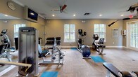 Located at 8134 Colonial Village Dr in Tampa, Florida, this well-equipped fitness center offers a comprehensive workout environment within a single-floor layout. The space features warm, neutral-toned walls complemented by crown molding, recessed lighting, and wood-style flooring throughout, creating a clean and inviting atmosphere.

The cardio section is anchored by a row of treadmills and elliptical trainers positioned along large multi-pane windows, offering pleasant views of the surrounding lush greenery and ample natural light. A ceiling fan overhead provides additional ventilation and comfort during workouts.

The strength training area is thoughtfully outfitted with a multi-station cable machine, a full dumbbell rack with a wall-length mirror, weight benches, and various resistance machines. TRX suspension trainers are mounted from ceiling brackets along one wall, accompanied by instructional exercise charts, making functional training easily accessible.

Additional amenities include a mounted flat-screen television, a wall-mounted AED defibrillator for safety, and a keypad-secured glass door providing direct outdoor access — offering views of what appears to be a pool area. Blue yoga mats are distributed across the floor to accommodate stretching and floor exercises. The space is bright, organized, and designed to support a wide range of fitness activities.
