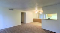 This single-floor residence offers a straightforward and functional layout designed for comfortable everyday living. The main living area is spacious and open, finished with wall-to-wall carpet flooring that adds warmth underfoot. Neutral-toned walls and a textured ceiling contribute to a clean, understated aesthetic throughout the space. A ceiling fan is centrally positioned, providing both lighting and air circulation to the room.

A defining feature of the living area is the partial-height counter that connects it to the kitchen, creating an open pass-through that enhances the sense of flow between the two spaces. The kitchen area is accented by a large mirrored panel, which reflects light and visually expands the room. A window adjacent to the mirror allows natural light to filter into the kitchen zone, brightening the interior.

A doorway off the main living area leads to the remainder of the unit, including additional rooms accessible via a short hallway. The overall layout is compact yet efficient, making good use of the available footprint. The consistent use of neutral tones, carpet, and practical fixtures throughout gives the space a cohesive, move-in-ready character suited to a range of living arrangements.