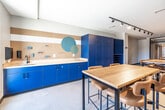 Located at 425 SW 11th St in Des Moines, Iowa, this single-floor collaborative workspace offers a thoughtfully designed environment suited for creative work, meetings, or group activities. The open layout accommodates multiple high-top wooden tables paired with tan leather-upholstered chairs, providing ample seating for group collaboration. A standout feature is the bold blue cabinetry wall, which includes both closed storage cabinets and an open cubby-style shelving unit, offering generous organizational capacity. Adjacent to the cabinetry, a pegboard workstation with a light-toned countertop and lower blue cabinets provides a dedicated hands-on workspace, accented by decorative teal circular graphics. Track lighting runs along the ceiling throughout the space, ensuring consistent, adjustable illumination. One wall displays a curated arrangement of clipboards in varying shades of blue, gray, and neutral tones, adding a functional yet artistic element to the interior. A full-height glass partition wall with a bold navy steel frame separates the space from an adjacent area, maintaining visual openness while defining boundaries. Natural light filters in through windows, complementing the light-toned floor. The overall aesthetic is modern and purposeful, balancing vibrant color accents with clean, neutral surfaces.