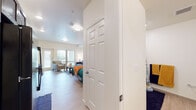 This single-floor studio unit offers a thoughtfully designed open-concept layout that seamlessly integrates the sleeping, living, and dining areas into one cohesive space. Light-toned hardwood-style flooring runs throughout, lending warmth and visual continuity to the interior.

The main living area is generously proportioned, accommodating both a sleeping area and a comfortable lounge space. Large floor-to-ceiling vertical blinds frame a sliding glass door that opens to a private balcony, filling the room with natural light and providing outdoor views. A wall-mounted split HVAC unit ensures year-round climate control.

The kitchen is efficiently laid out and features rich, dark walnut-toned cabinetry that extends to the ceiling for ample storage. Black appliances — including a range, dishwasher, and microwave — are paired with light countertops, creating a clean contrast. A stainless steel sink completes the functional workspace.

The bathroom is spacious and well-lit, showcasing matching dark wood cabinetry with an integrated vanity and sink, light-toned flooring, and a clean, minimalist aesthetic with grab bar accessibility features.

A six-panel interior door provides separation between the entry corridor and the main living space, adding a classic architectural detail. Overall, the unit presents a bright, modern, and functional living environment.