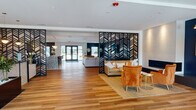This single-floor community clubhouse — identified as The Flats at Dorsett Ridge — delivers a polished, contemporary atmosphere from the moment you enter. The spacious lobby greets visitors with warm wood-look tile flooring, a sleek reception desk set against an artistic cityscape wall mural, and pendant lighting that anchors the open layout. Striking black laser-cut chevron partition screens serve as bold architectural dividers, elegantly defining each zone while maintaining a sense of openness throughout the floor.

The main lounge area features a linear fireplace set into a textured white accent wall, complemented by plush seating arrangements and a marble-topped coffee table. Adjacent to this is a lively game room with a large-screen TV mounted on a dramatic plaid feature wall, a shuffleboard table, and foosball — all centered around a generous sectional sofa. A branded area rug reinforces the community identity.

The dining and social area makes a striking impression with a deep navy wainscoted accent wall, gold sconce lighting, and a sculptural spur chandelier overhead. Mixed seating includes both a communal wood dining table and intimate bistro-style rounds, ideal for casual gatherings. A wet bar with navy cabinetry and decorative tile backsplash completes the entertaining amenities. Together, these spaces offer a cohesive, design-forward environment suited for both relaxation and community engagement.