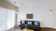 Located at 1005 W 5th Ave in Columbus, Ohio's sought-after Grandview Heights area, this single-floor space offers a thoughtfully designed contemporary interior. The living area immediately draws attention with its clean, minimalist aesthetic — bright white walls rise to tall ceilings, creating an open and airy atmosphere. Large windows dressed with sheer vertical panels invite abundant natural light to fill the room throughout the day. Rich dark hardwood flooring grounds the space and provides a striking contrast to the crisp white walls and ceiling. The living area is well-proportioned, comfortably accommodating modern furnishings while maintaining an uncluttered feel. A modern floor lamp and carefully curated décor, including colorful accent pillows and wall art featuring bold, linear patterns, add personality and warmth to the space. The overall design sensibility is modern and cohesive, balancing neutral tones with tasteful pops of color. The layout flows naturally, and the bright, welcoming ambiance makes the space feel both functional and inviting. Situated in a vibrant Columbus neighborhood with walkable access to local dining, shopping, and amenities, this space combines stylish interiors with an exceptional location.