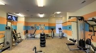 This fitness center offers a practical and energetic environment for exercise, distinguished by its bold horizontal color-blocked walls featuring alternating bands of orange and sky blue against a cream background. The room is single-story with a standard ceiling height, illuminated by a combination of recessed lighting and flush-mounted fluorescent fixtures that ensure consistent, bright lighting throughout the space.

The flooring consists of a durable, patterned tile that is well-suited for a high-traffic fitness environment. Equipment is thoughtfully arranged to maximize usable floor space. A freestanding dumbbell rack stocked with multiple weight sets serves as a central fixture, accompanied by a flat utility bench for strength training. Along the perimeter, a multi-function cable resistance machine provides options for a range of exercises, while two elliptical trainers offer low-impact cardiovascular workouts. A leg press machine adds to the strength training options available.

A window along one wall allows natural light to supplement the artificial lighting, contributing to an open and motivating atmosphere. The space is further accented by a piece of abstract wall art, adding a touch of visual interest to the functional environment.