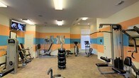 This fitness center offers a practical and energetic environment for exercise, distinguished by its bold horizontal color-blocked walls featuring alternating bands of orange and sky blue against a cream background. The room is single-story with a standard ceiling height, illuminated by a combination of recessed lighting and flush-mounted fluorescent fixtures that ensure consistent, bright lighting throughout the space.

The flooring consists of a durable, patterned tile that is well-suited for a high-traffic fitness environment. Equipment is thoughtfully arranged to maximize usable floor space. A freestanding dumbbell rack stocked with multiple weight sets serves as a central fixture, accompanied by a flat utility bench for strength training. Along the perimeter, a multi-function cable resistance machine provides options for a range of exercises, while two elliptical trainers offer low-impact cardiovascular workouts. A leg press machine adds to the strength training options available.

A window along one wall allows natural light to supplement the artificial lighting, contributing to an open and motivating atmosphere. The space is further accented by a piece of abstract wall art, adding a touch of visual interest to the functional environment.