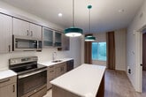 Located at 1550 Perimeter Pkwy NW in Huntsville, Alabama, this thoughtfully designed one-bedroom, one-bathroom unit makes excellent use of its compact footprint. The open-concept kitchen serves as the heart of the space, showcasing taupe-toned cabinetry with frosted glass upper panels, crisp white quartz countertops, and a full suite of stainless steel appliances including a refrigerator, range, microwave, and dishwasher. A central island with a white quartz surface provides additional prep space and casual seating, while two distinctive teal pendant lights add a bold pop of color overhead. Light wood-look flooring runs throughout, lending warmth and visual continuity across the space. The bedroom is comfortably sized, featuring a large window dressed with neutral linen-style drapes that filter in natural light. Two closets provide ample storage. The bathroom is finished in a clean, contemporary style with a wide vanity, white quartz countertop, dark cabinetry, and a backlit mirror. A recessed shelving niche and a stacked washer and dryer unit are conveniently integrated into the bathroom area, adding notable practicality. Throughout the unit, neutral tones, modern fixtures, and efficient layouts combine to create a functional yet stylish living environment.