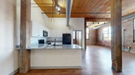 This 2-bedroom, 2-bathroom loft showcases a striking industrial aesthetic rooted in authentic architectural character. The open-concept living and kitchen area is anchored by soaring wood-plank ceilings with exposed timber beams and structural columns, complemented by polished hardwood-style flooring throughout. Exposed brick walls run along the perimeter, lending warmth and texture, while large industrial-style windows invite ample natural light into the expansive living space. Overhead, visible ductwork and track lighting add to the loft's distinctive urban character.

The kitchen features white cabinetry with granite countertops, a breakfast bar peninsula, and black appliances including a range, built-in microwave, and refrigerator. Both bathrooms are finished with granite vanity tops, white cabinetry, and tub-shower combinations. The generously sized primary bedroom offers carpeted flooring and access to a walk-in closet with wire shelving. The second bedroom similarly features carpet, exposed brick, and a closet with built-in shelving. Additional utility space and a dedicated closet round out the floor plan. The combination of raw industrial materials and updated finishes creates a cohesive, modern-loft atmosphere throughout.