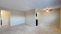 This single-floor space presents a clean, open layout designed for comfortable everyday living. The main living area is finished with neutral beige wall-to-wall carpeting and painted in soft, warm tones that create a bright and inviting atmosphere. The walls are smooth and painted in a light neutral color, complementing the overall simplicity of the space. White baseboards line the perimeter, adding a subtle finishing touch.

A partial dividing wall separates the living area from the adjacent kitchen zone, providing a degree of definition between the two spaces while maintaining an open feel. The kitchen area features dark cabinetry visible above and a dark appliance, likely a refrigerator, positioned near the transition point. A ceiling fan with a light fixture is installed overhead in the kitchen area, offering both ventilation and ambient lighting. Electrical outlets are distributed along the walls at standard intervals, ensuring practical functionality throughout. The overall ambiance is calm and understated, with a neutral palette that provides a versatile backdrop suitable for a variety of furnishing styles.