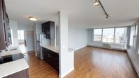 This well-appointed single-floor space showcases a thoughtfully designed open-concept layout that seamlessly connects the kitchen and living areas. The kitchen is outfitted with rich dark cabinetry, a stainless steel refrigerator, and light-toned countertops that extend into a convenient breakfast bar peninsula — an ideal spot for casual dining or entertaining. A structural column adds architectural character while subtly defining the transition between the kitchen and the expansive living area. Warm hardwood flooring flows throughout the entire space, lending a cohesive and inviting aesthetic. The living area is generously proportioned and bathed in natural light streaming through large floor-to-ceiling windows, which frame sweeping views of the surrounding cityscape. Track lighting in the living area complements the natural light, offering flexible illumination options. A baseboard heating unit and wall-mounted air conditioning ensure year-round comfort. The neutral grey and white wall palette creates a clean, versatile backdrop, making the space easy to personalize. Overall, the unit delivers a bright, modern, and functional living environment with a strong urban character.
