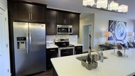 This well-appointed single-floor space showcases a thoughtfully designed interior with a strong emphasis on modern aesthetics and functionality. The kitchen serves as a central highlight, featuring rich dark espresso cabinetry that extends to the ceiling, offering generous storage capacity. Stainless steel appliances, including a French door refrigerator, an over-the-range microwave, and an electric range, provide a sleek, cohesive look throughout the cooking area. The light-toned stone countertops offer a striking contrast against the dark cabinetry, creating a sophisticated and contemporary feel. A kitchen island with an undermount sink adds both workspace and a casual gathering spot, enhancing the room's functionality. The neutral light gray walls complement the overall color palette, while the open layout promotes a sense of spaciousness and easy flow between areas. A glimpse into the adjoining spaces reveals consistent modern finishes and an airy ambiance. Natural and ambient lighting work together to keep the space bright and inviting. Overall, the space combines practical design with stylish details, resulting in a comfortable and visually appealing environment.