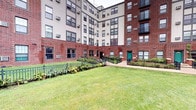 This residential building presents a striking exterior defined by a combination of rich red brick at the base and cream-colored siding on the upper floors, creating a layered architectural aesthetic. The structure rises several stories and is arranged in a U-shaped configuration, enclosing a central courtyard that adds a sense of community and openness to the property.

The courtyard is a notable feature, offering a well-maintained lawn bordered by ornamental plantings, including broad-leafed tropical-style foliage that provides visual interest against the iron fencing lining the perimeter. Pathways lead to multiple entry points, including a distinctive teal-painted door that serves as a welcoming focal point along the ground-level facade.

Windows are evenly distributed across all floors, providing natural light throughout the building, and wall-mounted air conditioning units are visible, indicating individual climate control for each unit. The surrounding outdoor grounds extend beyond the building, featuring mature trees, curved walkways, and expansive grassy areas that contribute to a park-like setting. The overall ambiance is one of established urban living, combining functional design with accessible green spaces.