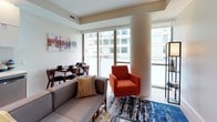Located at 301 M Street, Unit 410 in Washington, DC, this thoughtfully designed residence offers a contemporary urban living experience across a single floor. The open-concept kitchen is outfitted with stainless steel appliances, including a full-size refrigerator, electric range with oven, built-in microwave, and dishwasher. Rich walnut-toned lower cabinetry is paired with crisp white upper cabinets and a clean white countertop, creating a modern, functional workspace. Light wood-style flooring flows from the kitchen into the living and dining area, where floor-to-ceiling sliding glass doors flood the space with natural light and offer views of the surrounding cityscape. A rounded architectural column adds a distinctive design touch to the living area. The bedroom features plush carpeting, bold accent walls in a warm yellow tone, and a built-in desk nook — ideal for a home workspace. Decorative wall art and ample room for furniture make it a comfortable retreat. An adjacent bathroom is finished with light tile flooring, a white undermount sink set in dark wood cabinetry, chrome fixtures, and a separate water closet for added privacy. The overall aesthetic blends modern design with warm, vibrant accents throughout.