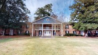 Situated in Charleston, South Carolina, this property at 1840 Carriage Ln offers a charming blend of traditional Southern architecture and timeless curb appeal. The exterior is defined by classic red brick construction, complemented by a striking covered front porch supported by white columns and a white balustrade — a hallmark of Lowcountry-influenced design. A decorative circular window accent at the roofline adds a distinctive architectural touch, while blue-gray siding on the upper gable lends subtle contrast to the warm brick facade. Double white entry doors provide a welcoming focal point at the front of the building. Mature trees, including towering pines and broad-canopied hardwoods, frame the property and provide natural shade, enhancing the serene, established character of the setting. Black shutters on the windows create a crisp, classic aesthetic consistent throughout the facade. The expansive front lawn, with its well-maintained open grassy area, contributes to the sense of space and tranquility. Nestled within a quiet area of West Ashley, this property benefits from its peaceful residential surroundings while remaining conveniently accessible to the broader Charleston area.