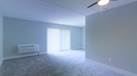 This single-floor space offers a clean, functional layout designed with simplicity and practicality in mind. The main living area is finished with wall-to-wall carpeting in a neutral gray tone, providing a soft and comfortable underfoot feel throughout. The walls are painted in a light, cool gray-white hue, creating a calm and airy atmosphere that enhances the sense of openness within the space.

A large window dressed with full-length vertical blinds serves as the focal point of the main room, allowing an abundance of natural light to filter in while offering flexible light control and privacy. A ceiling fan is installed overhead, contributing both to air circulation and to the overall functional character of the space.

A wall-mounted air conditioning unit is positioned along one wall, ensuring climate control comfort year-round. The ceiling features a paneled design, adding a subtle architectural detail to the otherwise streamlined interior.

The space presents a straightforward, move-in-ready environment well-suited for a variety of uses. Its neutral palette and open layout offer a versatile foundation that can be easily adapted and personalized to suit a range of furnishing styles and functional needs.