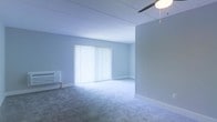 This single-floor space offers a clean, functional layout designed with simplicity and practicality in mind. The main living area is finished with wall-to-wall carpeting in a neutral gray tone, providing a soft and comfortable underfoot feel throughout. The walls are painted in a light, cool gray-white hue, creating a calm and airy atmosphere that enhances the sense of openness within the space.

A large window dressed with full-length vertical blinds serves as the focal point of the main room, allowing an abundance of natural light to filter in while offering flexible light control and privacy. A ceiling fan is installed overhead, contributing both to air circulation and to the overall functional character of the space.

A wall-mounted air conditioning unit is positioned along one wall, ensuring climate control comfort year-round. The ceiling features a paneled design, adding a subtle architectural detail to the otherwise streamlined interior.

The space presents a straightforward, move-in-ready environment well-suited for a variety of uses. Its neutral palette and open layout offer a versatile foundation that can be easily adapted and personalized to suit a range of furnishing styles and functional needs.