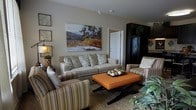 This single-story residence presents a cohesive, open-concept layout that seamlessly connects the main living areas. A welcoming entryway with light hardwood-style flooring leads directly into the heart of the home, where the dining area, kitchen, and living room flow naturally together.

The kitchen is outfitted with rich, dark espresso cabinetry that extends to the upper walls, offering generous storage. A tile backsplash adds texture behind the countertops, and the space is well-lit with overhead lighting. Adjacent to the kitchen, a dining area accommodates a dark wood table and chairs set atop a patterned area rug, creating a defined yet open dining space.

The living room is warm and inviting, anchored by a neutral-toned sofa paired with patterned accent chairs and an orange upholstered ottoman that introduces a bold pop of color. A large landscape painting serves as a focal point above the sofa, while complementary wall décor and greenery add depth and personality throughout. The consistent flooring and neutral wall tones unify the entire space, giving the home a cohesive, well-curated ambiance.