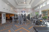 This contemporary fitness center is designed to offer a comprehensive workout experience within a single, open-plan floor. The space immediately impresses with soaring ceilings and an abundance of natural light streaming through large floor-to-ceiling windows that frame views of a lush outdoor pergola and surrounding greenery, creating an energizing atmosphere throughout.

The interior design is anchored by a distinctive herringbone-patterned carpet in grey and yellow tones, lending the space a dynamic, modern aesthetic. Bold, abstract line artwork in orange and red sweeps across the white walls, adding an artistic flair that complements the energetic purpose of the facility.

The main cardio area features a well-curated row of treadmills, elliptical trainers, and cycling machines, all positioned to take advantage of the outdoor views. Floor-to-ceiling mirrors expand the sense of space and allow users to monitor their form. A dedicated strength training zone offers a comprehensive cable machine system alongside a full rack of dumbbells and adjustable weight benches.

A separate functional training area is equipped with an orange and black multi-purpose rig loaded with kettlebells, medicine balls, resistance bands, and suspension trainers. A heavy punching bag and wall-mounted training pads make this corner ideal for combat fitness and high-intensity interval training. The fitness center rules are clearly posted, and the overall layout ensures ample room for movement and exercise variety.