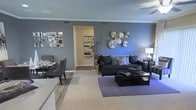 Located at 1250 S. Dayton Ct in Denver, Colorado, this single-floor residence offers a thoughtfully designed living space that balances style and comfort. The open-concept living and dining area serves as the heart of the home, featuring warm, neutral carpeting that complements the bold, steel-blue accent walls. Decorative wall art, including geometric mirror arrangements and metallic sculptural pieces, adds a contemporary flair to the space. The living area is generously proportioned, accommodating a full sectional sofa, a square coffee table, and an area rug, all anchored beneath a ceiling fan that enhances air circulation and comfort. A dining nook sits adjacent to the living space, seamlessly integrating the two areas for an open, flowing feel. A central hallway connects to additional rooms, maintaining a logical and efficient floor plan throughout. Crown molding along the ceilings provides a polished architectural finish, while multiple overhead vents suggest a well-maintained climate control system. The overall ambiance is modern and inviting, with cohesive décor and a neutral foundation that makes the space feel both spacious and welcoming.