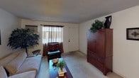 This single-floor residence offers a practical and comfortable layout designed for everyday living. The living room serves as the heart of the space, featuring wall-to-wall carpeting, a textured ceiling, and a wall-mounted air conditioning unit. A large window dressed with dark curtains allows natural light to filter in, brightening the room. The space comfortably accommodates a plush sofa, a leather accent chair with ottoman, a coffee table, and a large wooden armoire, making it both functional and inviting. Framed artwork and decorative indoor plants add warmth and personality to the walls and corners.

The kitchen is efficiently designed, featuring warm wood-toned upper cabinetry, a coil-burner electric range, a refrigerator, and a sink — all thoughtfully arranged to maximize workflow in a compact footprint. A dining area adjacent to the kitchen provides space for a table and seating, making mealtimes convenient and accessible.

A utility area off one of the hallways houses a full-size water heater along with shelving, offering practical storage solutions. The overall layout, as seen in the dollhouse view, flows in a connected and logical manner, making excellent use of the available space throughout this single-level home.