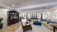 This expansive single-floor space is thoughtfully designed to serve as a welcoming communal gathering area. The layout encompasses multiple distinct lounge zones, a fully equipped kitchen, and several transition areas, all unified by a cohesive navy blue and warm neutral color scheme throughout.

The main lounge area is generously proportioned, featuring tray ceilings with recessed lighting and large windows dressed in bold navy patterned drapes that allow abundant natural light to fill the space. Seating arrangements include classic upholstered sofas, wooden armchairs, and a striking navy blue ottoman, complemented by dark wood furniture and a built-in bookcase along one wall.

Additional seating alcoves offer more intimate settings, with large windows providing outdoor views and continued use of the signature navy drapery. Stone tile flooring transitions seamlessly between areas, while plush area rugs define individual seating zones.

The kitchen is well-appointed with rich cherry wood cabinetry, granite countertops, stainless steel appliances including a gas range and French door refrigerator, and a central island providing ample workspace. Throughout the space, warm beige walls, tasteful artwork, and lush potted plants contribute to an inviting and polished ambiance.