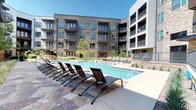 This Dallas property is defined by its striking courtyard-centered design, where a large, resort-style swimming pool serves as the focal point of the community. The surrounding four-story building features a contemporary architectural aesthetic that blends warm wood-tone siding, stacked stone cladding, and crisp white stucco panels, creating a visually dynamic exterior. Teal-framed windows and private balconies with sleek metal railings extend outward from multiple levels, offering residents direct views of the pool and landscaped grounds below.

The pool deck is finished with natural stone pavers and is generously appointed with rows of adjustable lounge chairs, providing ample space for sunbathing and relaxation. The pool itself is expansive, featuring clear blue water with a shallow wading entry and stainless steel handrails. Lush native landscaping, including ornamental grasses and small trees, is thoughtfully integrated throughout the courtyard, softening the hardscape and adding greenery to the environment. The open-sky courtyard layout ensures abundant natural sunlight reaches the pool area throughout the day, enhancing the overall outdoor living experience.