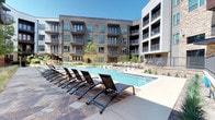 This Dallas property is defined by its striking courtyard-centered design, where a large, resort-style swimming pool serves as the focal point of the community. The surrounding four-story building features a contemporary architectural aesthetic that blends warm wood-tone siding, stacked stone cladding, and crisp white stucco panels, creating a visually dynamic exterior. Teal-framed windows and private balconies with sleek metal railings extend outward from multiple levels, offering residents direct views of the pool and landscaped grounds below.

The pool deck is finished with natural stone pavers and is generously appointed with rows of adjustable lounge chairs, providing ample space for sunbathing and relaxation. The pool itself is expansive, featuring clear blue water with a shallow wading entry and stainless steel handrails. Lush native landscaping, including ornamental grasses and small trees, is thoughtfully integrated throughout the courtyard, softening the hardscape and adding greenery to the environment. The open-sky courtyard layout ensures abundant natural sunlight reaches the pool area throughout the day, enhancing the overall outdoor living experience.