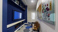 This dedicated game room is a thoughtfully designed entertainment space that blends bold style with functional layout. The focal point of the room is a striking navy blue built-in wall unit, which houses a large flat-screen television, open display shelves showcasing trophies and collectibles, and lower cabinet storage with woven basket drawers — offering both visual impact and practical organization. A continuous white ledge below the screen provides a convenient surface for media components and accessories. Opposite the entertainment wall, a row of vintage-style wooden stadium seats lines the room, giving the space a fun, arena-inspired character perfect for gaming and movie watching. The flooring features a geometric hexagonal tile pattern in blue, white, and gray tones that adds a dynamic, modern energy to the room. Large-scale pop-art and entertainment-themed wall prints, including a prominent Star Wars graphic, adorn the neutral gray walls, enhancing the playful atmosphere. A white paneled door with glass detailing at the far end allows natural light to filter in, brightening the space. Recessed ceiling lighting and crown molding add a polished, finished quality to this entertaining haven.