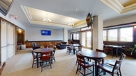 Located at 755 E Broad St in Athens, GA, this well-appointed single-floor facility offers a thoughtfully designed layout suited for gatherings and communal use. The centerpiece of the space is a large combined dining and living room, characterized by an elegant tray ceiling adorned with classic chandeliers and recessed lighting. Multiple wall-mounted televisions are strategically positioned throughout, enhancing the room's functionality. Dark wood round dining tables paired with cushioned chairs are arranged throughout the carpeted space, encouraging both formal and casual interaction. A built-in wooden cabinetry unit with dark granite countertops runs along one wall, adding a refined, traditional aesthetic. Plush leather sofas and a warm wooden booth-style seating nook provide additional lounge areas. Large glass doors open to an exterior view, flooding the space with natural light. The kitchen is outfitted with ample wood cabinetry, generous counter space, and stainless steel appliances, including a range and small appliances. The six bathrooms feature neutral travertine-style tile flooring and wainscoting, with accessibility-friendly fixtures including grab bars. A connecting hallway ties the spaces together, offering a seamless and functional flow throughout the facility.