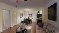 This single-floor residence showcases an open-concept layout that seamlessly connects the living, dining, and kitchen areas into one cohesive, flowing space. Warm hardwood flooring runs throughout, lending a sense of continuity and warmth to the interior.

The living area is anchored by a tufted sofa and a sleek oval glass coffee table, accented by a decorative potted plant that adds a natural touch. A ceiling fan overhead provides both function and style. Crown molding along the upper walls adds a refined architectural detail.

The dining area transitions naturally from the living space, centered around a round dining table with dark wood chairs and illuminated by a chandelier with multiple lights, creating an inviting atmosphere for meals. A decorative framed mirror on the wall enhances the sense of space.

The kitchen, visible from the main living area, features dark wood cabinetry, stone countertops, and modern appliances. Its open sightline to the dining and living areas reinforces the home's open, social design.

Overall, the space presents a warm, well-appointed interior with a cohesive color palette, balanced lighting, and thoughtfully arranged furnishings.
