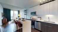 This single-floor space showcases a contemporary open-concept layout that seamlessly integrates the kitchen and living areas. The kitchen is designed with clean lines, featuring floor-to-ceiling white cabinetry that maximizes vertical storage space. Stainless steel appliances, including a range, microwave, and dishwasher, are complemented by a crisp white countertop that extends into a peninsula-style breakfast bar. The bar seating, fitted with warm brown leather-toned stools, provides a casual dining and entertaining option.

Dark wood-grain lower cabinetry contrasts elegantly with the white upper units, adding visual depth to the kitchen. An industrial-style open shelving unit with a black metal frame serves as a stylish transition between the kitchen and living area, offering both display and storage functionality.

The living space benefits from generous natural light streaming through large windows, dressed with teal curtains that add a pop of color. City views are visible through the windows, enhancing the urban character of the space. Dark hardwood flooring runs throughout, grounding the light-toned walls and creating a warm, cohesive aesthetic. Overhead, track-style lighting provides targeted illumination across both functional zones.