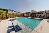 This outdoor community amenity area centers around a generously sized swimming pool with crystal-clear blue water, designed to accommodate both leisure and lap swimming. The pool features built-in water jets and dedicated in-water lounge chairs along a shallow ledge entry, providing a resort-style experience for residents. A broad, light-colored concrete sundeck surrounds the pool on all sides, lined with multiple chaise lounges and seating arrangements to accommodate sunbathing and relaxation throughout the day.

The pool area is enclosed by a black wrought-iron fence, providing both security and an open, airy feel. Classic lantern-style lamp posts are positioned throughout the deck, adding architectural charm and functional lighting. Nearby, a pergola structure and shaded umbrella seating offer additional gathering spots.

The surrounding landscape features newly planted trees and manicured greenery, set against a natural hillside backdrop. Modern farmhouse-style residential buildings with white siding and dark rooflines frame the perimeter, contributing to a cohesive and well-planned community aesthetic. The overall ambiance is bright, open, and inviting, well-suited for outdoor recreation under ample natural sunlight.