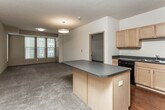 This single-floor unit is thoughtfully designed with an open-concept layout that seamlessly connects the kitchen and living area. The kitchen is well-appointed with light maple cabinetry, dark laminate countertops, and black appliances, including a range, refrigerator, and dishwasher. A central island provides additional prep and storage space. Rich hardwood flooring runs throughout the kitchen, while plush carpet covers the living areas and bedrooms. The open living space is generously sized, offering room for multiple furniture arrangements and benefiting from natural light through several windows fitted with blinds. A dedicated in-unit laundry area with a stacked washer and dryer adds everyday convenience. The unit includes two bedrooms, each carpeted and equipped with windows that overlook the surrounding building exterior. Two bathrooms serve the unit: one features a tub-shower combination with neutral tile surround, and the other includes a vanity with ample under-sink storage and a large mirror. Textured ceilings and consistent neutral wall tones create a cohesive, understated ambiance throughout the space.