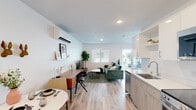 This thoughtfully designed single-floor studio offers a clean, contemporary aesthetic throughout. The open-concept layout seamlessly connects the kitchen, dining, living, and sleeping areas, creating an efficient and cohesive living environment bathed in natural light from multiple windows and a sliding glass door.

The kitchen is well-appointed with white upper cabinetry, light wood-toned lower cabinets, white quartz countertops, a stainless steel sink, electric range, built-in microwave, and a full-size refrigerator — providing a complete set of appliances for everyday cooking.

The main living area flows naturally into the sleeping space, with light-toned hardwood-style flooring extending throughout. A dedicated workspace with a mid-century modern desk adds functional versatility. The sleeping area accommodates a full-size bed alongside a green tufted sofa, and features a sliding glass door opening directly to a private outdoor patio.

The bathroom is clean and minimal, finished with light flooring and a shower/tub combination. A wall-mounted mini-split HVAC unit provides climate control. Recessed lighting and crisp white walls contribute to a bright, airy ambiance throughout the entire unit.