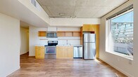 Located at 11011 NE Ninth St in Bellevue, WA, this three-bedroom, two-bathroom unit offers a well-organized single-floor layout that makes efficient use of its space. The architectural design incorporates exposed concrete ceilings, which lend an industrial-modern character throughout the unit, complemented by warm-toned wood-style flooring in the main living areas and neutral carpeting in the bedrooms.

The kitchen is thoughtfully equipped with stainless steel appliances, including a refrigerator, electric range, microwave, and dishwasher. Warm-toned wood cabinetry with open shelving provides both functional storage and visual appeal, while a white countertop offers ample workspace. Track lighting above the kitchen highlights the soaring exposed concrete ceiling.

The bedrooms are carpeted and benefit from generously sized windows that frame views of Bellevue's urban skyline, filling rooms with abundant natural light. The primary bedroom includes a sliding-door closet, while a separate dedicated closet adds further storage capacity.

The two bathrooms feature clean, functional designs. One includes a walk-in glass-door shower with chrome fixtures, while the other offers a warm-toned wood vanity with a white countertop. Both maintain a consistent, neutral aesthetic throughout.