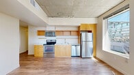 Located at 11011 NE Ninth St in Bellevue, WA, this three-bedroom, two-bathroom unit offers a well-organized single-floor layout that makes efficient use of its space. The architectural design incorporates exposed concrete ceilings, which lend an industrial-modern character throughout the unit, complemented by warm-toned wood-style flooring in the main living areas and neutral carpeting in the bedrooms.

The kitchen is thoughtfully equipped with stainless steel appliances, including a refrigerator, electric range, microwave, and dishwasher. Warm-toned wood cabinetry with open shelving provides both functional storage and visual appeal, while a white countertop offers ample workspace. Track lighting above the kitchen highlights the soaring exposed concrete ceiling.

The bedrooms are carpeted and benefit from generously sized windows that frame views of Bellevue's urban skyline, filling rooms with abundant natural light. The primary bedroom includes a sliding-door closet, while a separate dedicated closet adds further storage capacity.

The two bathrooms feature clean, functional designs. One includes a walk-in glass-door shower with chrome fixtures, while the other offers a warm-toned wood vanity with a white countertop. Both maintain a consistent, neutral aesthetic throughout.