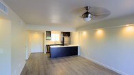 This single-floor space showcases a clean, modern design centered around an open-concept layout that seamlessly connects the kitchen and living areas. The kitchen is a standout feature, equipped with dark cabinetry, a large island with a bright white countertop, a stainless steel refrigerator, a dishwasher, and a built-in cooktop. The island also functions as a casual dining or prep space, making it both practical and visually striking against the light hardwood-style flooring that extends throughout the unit.

The living area is open and airy, with recessed lighting along a dropped ceiling section adding a layered architectural detail. A ceiling fan with a brushed nickel finish provides both function and a contemporary accent. Large sliding glass doors flood the space with natural light and open directly to an outdoor area, enhancing the sense of openness and connectivity to the exterior.

The walls are finished in neutral tones, contributing to the space's bright, minimalist ambiance. A private entry door is also visible, providing a clear sense of the unit's layout and flow. Overall, the space offers a well-appointed, functional environment with modern finishes and a thoughtfully designed open floor plan.