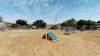 Located in Abilene, Texas, this single-level outdoor community space offers a range of practical amenities designed to serve residents of the surrounding multi-building complex. The grounds are characterized by open, grassy areas enclosed by chain-link fencing, with mature trees providing natural shade throughout the property.

A standout feature is the dedicated dog park, a generously sized fenced area equipped with agility equipment including ramps and A-frame obstacles. The park is set against a backdrop of open sky and surrounding greenery, offering pets and their owners a functional and spacious outdoor area for activity.

A covered mailbox station serves the community with an extensive array of individual mailboxes in yellow and silver, organized across two rows under a wood-framed, pitched roof structure. The open-sided design allows for natural ventilation while providing shelter from the elements. The station is conveniently accessible from the adjacent parking area and walkways.

The broader grounds include paved parking areas, concrete walkways, picnic seating visible in the distance, and unobstructed views of the surrounding landscape. The overall character of the space is utilitarian and community-focused, providing residents with essential outdoor amenities in a spacious, open-air setting typical of West Texas developments.