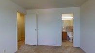 This single-floor space is straightforward in its layout, offering a functional and practical design. The main room features wall-to-wall carpeting in a neutral beige tone, contributing to a quiet and comfortable atmosphere. The walls are finished in a light gray-blue hue, keeping the space feeling clean and understated.

A standard closet with a white paneled door is positioned along one wall, providing convenient built-in storage. An additional door leads to a neighboring room or hallway, suggesting easy connectivity within the floor plan.

Directly accessible from the main room is an en-suite bathroom, visible through an open doorway. The bathroom is equipped with a single-sink vanity featuring warm wood-toned cabinetry, a large mirror with overhead strip lighting that ensures well-lit functionality, and a toilet. The flooring transitions to a patterned tile, contrasting with the carpeted main room. The bathroom also appears to include a tiled tub or shower enclosure along the far wall.

Overall, the space presents a modest, no-frills design that prioritizes practicality, making it suitable for everyday residential use.